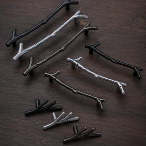 May include: A collection of cabinet hardware pulls and knobs shaped like tree branches. The set includes various lengths and finishes, including matte black, brushed silver, and dark metallic gray, each featuring a textured, organic twig design.