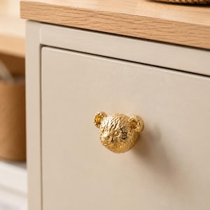 May include: A gold-colored metal cabinet knob shaped like a leopard head with textured spots and rounded ears, mounted on a smooth, light beige drawer front with a light wood-toned top surface visible above the drawer.