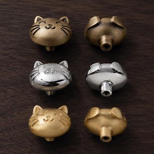 May include: A set of six cat-shaped cabinet knobs arranged in two columns. The knobs feature a smiling feline face with pointed ears and whiskers. The set includes three knobs with a brushed brass finish, two with a polished silver finish, and one matte gold.