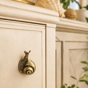 May include: A brass-toned metal cabinet knob shaped like a snail with a textured shell and body. The snail is mounted on a light beige wooden cabinet door with a vertical grain. The hardware features a metallic finish with dark highlights.