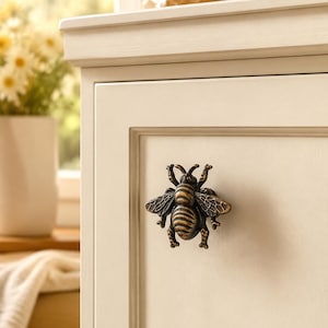May include: A bronze-toned metal cabinet knob shaped like a bee with detailed wings and a segmented body. The bee knob is mounted on a flat, off-white cabinet door panel, featuring a dark, weathered finish that highlights the insect's texture.