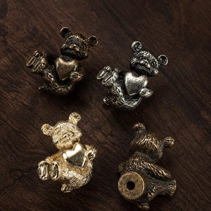 Puede incluir: Cuatro pequeñas figuras de oso de metal, cada una sosteniendo un corazón. Tres osos se muestran de frente con detalles de pelaje texturizado y corazones lisos, mientras que un oso se muestra de espaldas, revelando un orificio circular en la base.