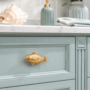 May include: A gold-toned metal cabinet knob shaped like a fish with detailed scales and fins, mounted on a light blue-green drawer front with a paneled frame. The drawer features a smooth, matte finish and a rectangular design with decorative molding.