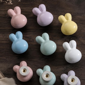 May include: A collection of nine ceramic cabinet knobs shaped like rabbit heads in pastel colours including pink, lavender, yellow, blue, mint green, and white. Each knob features two upright ears and a smooth, glossy finish with a metal screw insert.