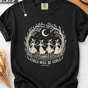 May include: A black short-sleeve t-shirt featuring a beige graphic of four figures in pointed hats dancing in a circle under a crescent moon and tree branches. The text "GIRLS WILL BE GIRLS" is printed in a curved beige font below the illustration.