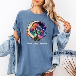 Può includere: Una t-shirt blu a girocollo con la grafica di un albero colorato all'interno di una luna crescente e un cielo stellato. Sotto la grafica, il testo recita FREEDOM • DIGNITY • TRANQUILITY. La t-shirt è indossata con una giacca di jeans blu.
