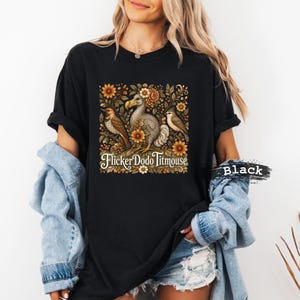 May include: A black t-shirt featuring a central graphic of a dodo, a flicker, and a titmouse surrounded by orange and brown flowers. The text "FlickerDodoTitmouse" is printed in a white serif font below the birds. The word "Black" is visible.