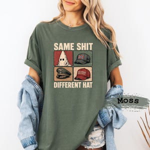 Puede incluir: Una camiseta de manga corta color verde musgo con un estampado gráfico que incluye el texto SAME SHIT DIFFERENT HAT. El diseño presenta cuatro paneles cuadrados que muestran una capucha blanca, una gorra con bandera, una gorra militar y una gorra roja.