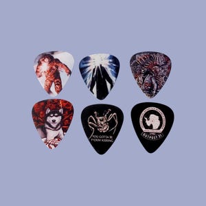 May include: Six guitar picks with different designs. The picks feature images of a creature from the movie "The Thing", a wolf, a spider, a creature from the movie "The Thing", a creature from the movie "The Thing", and a logo for "Outpost 31".
