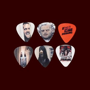 May include: Six guitar picks featuring images from the movie "The Shining". The picks are white with red and black accents. The images include Jack Nicholson, Danny Lloyd, Shelley Duvall, and the Grady twins.