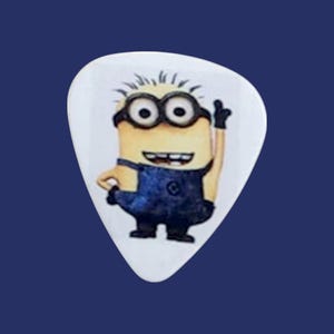 May include: A white guitar pick with a cartoon image of a yellow minion wearing blue overalls and goggles. The minion is smiling and has one arm raised.