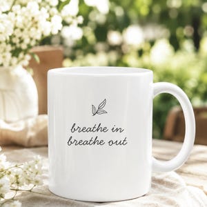 May include: A white ceramic mug featuring a black line drawing of a small leaf sprig above the black text "breathe in breathe out" in a simple script font. The mug has a smooth finish, a cylindrical body, and a curved handle on the right side.