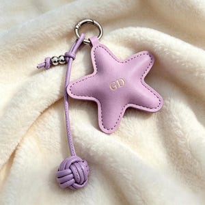 May include: A lavender star-shaped leather keychain featuring the gold-embossed letters GD in the center. The charm is attached to a silver metal ring with a matching lavender leather cord and a decorative knotted leather ball at the end.