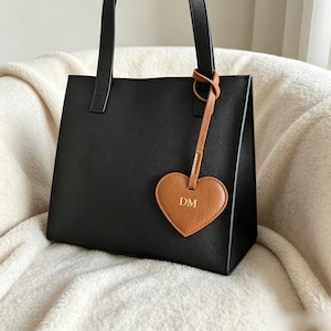 May include: A structured black tote bag with two top handles, featuring a tan heart-shaped leather luggage tag attached to one handle. The tag is embossed with the gold-colored letters DM in the center, providing a contrast to the black bag.