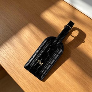 以下が含まれることがあります： 光沢のあるクロコダイル調の型押しが施された黒いラゲッジタグ。ゴールドトーンの金属製バックルストラップが付いています。中央にはゴールドカラーのモノグラム文字「FM」がプリントされており、旅行用バッグの明確な識別マーカーとして機能します。