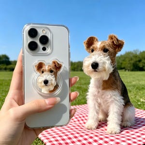May include: A clear smartphone case featuring a circular pop-out grip with a printed image of a wire-haired terrier dog. The dog has a white muzzle, brown ears, and a tan and white coat. The phone case is held up against a background of a dog.