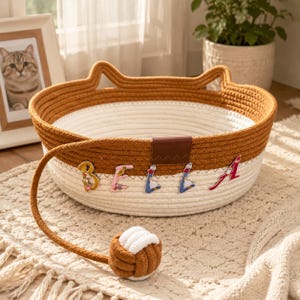 Puede incluir: Una cesta redonda de cuerda tejida en dos tonos con una sección superior marrón con bordes en forma de orejas de gato y una sección inferior blanca. El nombre BELLA está escrito con letras decorativas de motivos florales. Incluye un juguete de cuerda.