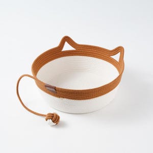 Pode incluir: Uma cesta redonda de corda trançada com interior branco e borda externa marrom, apresentando duas formas pontiagudas de orelhas de gato no topo. Um cordão longo estende-se da lateral da cesta, terminando em uma pequena bola nó de corda.
