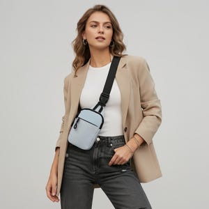 May include: A light blue crossbody bag with a black zipper and a black adjustable strap. The bag features a rectangular shape with rounded corners and a front pocket, worn over a white t-shirt, a beige blazer, and dark gray denim jeans.