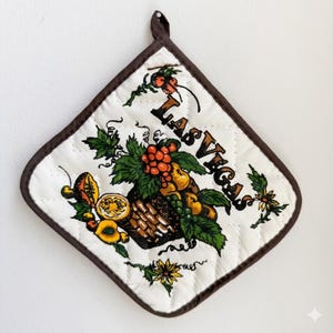 May include: A diamond-shaped quilted pot holder with a brown fabric border and a hanging loop. The white fabric features a colorful graphic of a fruit basket with green leaves and the text LAS VEGAS printed in a bold, dark brown font.