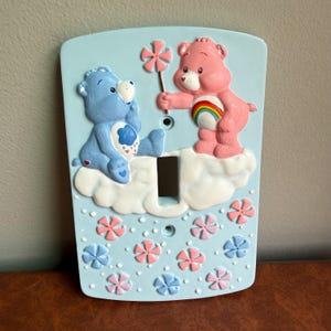 May include: A light blue plastic light switch plate featuring a raised relief design of a blue bear and a pink bear with a rainbow belly badge. The bears sit on a white cloud, and the lower section is decorated with small pink and blue flowers.