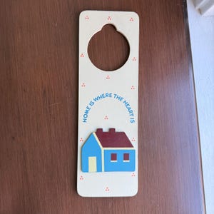 May include: A cream-colored wooden door hanger featuring a circular cutout at the top. The text "HOME IS WHERE THE HEART IS" is printed in blue above a three-dimensional wooden house icon with a blue exterior, a dark red roof, and a yellow door.