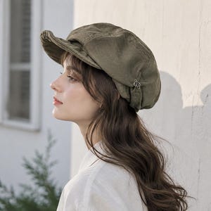 Può includere: Un berretto in stile newsboy color verde oliva con visiera strutturata e corona morbida e arrotondata. Il cappello è impreziosito da un piccolo ciondolo metallico color argento a forma di nodo complesso sul lato, come dettaglio decorativo.