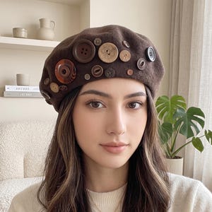 May include: A brown felt beret adorned with an assortment of various sized buttons in shades of brown, tan, and black. The buttons are arranged across the front of the hat, featuring different textures, patterns, and materials including wood and plastic.