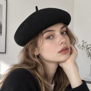 May include: A solid black knit beret featuring a ribbed texture and a small, centered top knot. The hat is constructed from a soft, thick yarn material with a rounded, structured shape that sits comfortably on the head, providing a classic look.