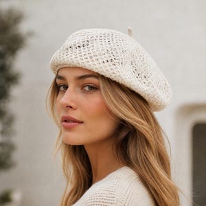 May include: A cream-colored beret crafted from a lightweight, open-knit crochet material. The hat features a circular, structured crown with a small, centered knot at the top and a soft, flexible brim that sits comfortably around the head.