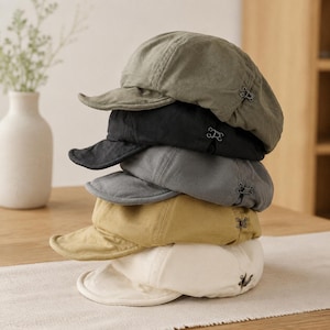 May include: A stack of five baseball caps in olive green, black, charcoal grey, mustard yellow, and off-white. Each cap features a short, curved brim and a small, metallic hook-and-eye fastener detail on the side panel near the back of the crown.