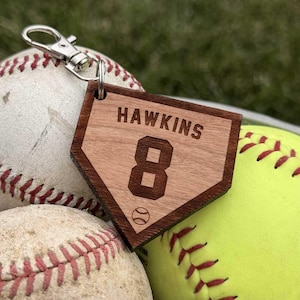 Könnte beinhalten: Ein hölzerner Schlüsselanhänger in Form einer Baseball-Homeplate mit der Gravur HAWKINS und der Nummer 8 sowie einer kleinen Baseball-Grafik. Der Anhänger ist an einem silberfarbenen Karabinerhaken befestigt und liegt auf zwei Baseballs.