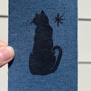 May include: A rectangular blue denim fabric patch featuring a black silhouette of a sitting cat facing left. Above the cat's head is a small black star-shaped icon. The patch has a visible twill weave texture throughout the blue material.