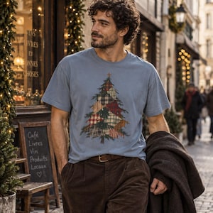 May include: A dusty blue short-sleeve t-shirt featuring a graphic of a Christmas tree composed of various plaid and tartan fabric patterns in shades of red, green, brown, and cream, with a small star at the top of the tree design.