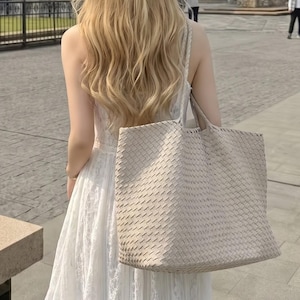 May include: A large, beige tote bag featuring an intricate woven pattern across its entire surface. The bag has two shoulder straps and is carried on the side, showcasing a structured rectangular shape with a consistent, uniform texture throughout.