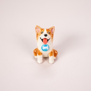May include: A small ceramic figurine of a Corgi dog with brown and white fur. The dog is sitting with its tongue out and wearing a blue circular collar tag featuring a white bone graphic. The figurine has a glossy finish and painted details.