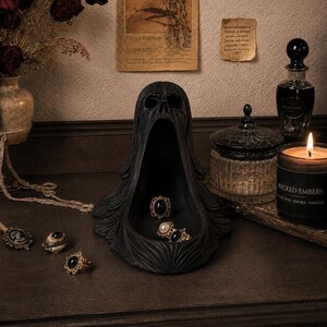 May include: A black, hollow, skull-shaped sculpture with a wide, open mouth serving as a tray. The sculpture features a hooded, draped appearance and is positioned on a surface with several rings placed inside the open mouth area.