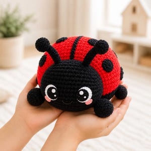 May include: A round, crocheted ladybird plush toy featuring a bright red back with black circular spots and a black head. The toy has two antennae with rounded tips, large black eyes with white highlights, and small pink cheeks stitched onto the face.