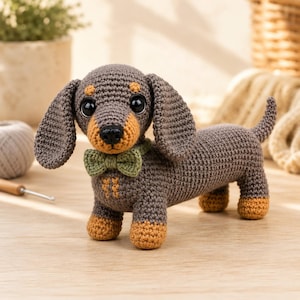 May include: A crocheted dachshund toy featuring a dark brown body, tan paws, and a tan muzzle. The dog has large black plastic eyes, a black nose, and wears a small olive green crocheted bow tie around its neck. The texture is consistent throughout.