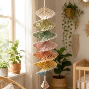 Könnte beinhalten: Ein hängendes Spiral-Mobile aus gehäkelten Spitzenscheiben in einem Farbverlauf aus Creme, Salbeigrün, zartem Rosa, Pfirsich, Gelb und Hellblau. Das Mobile ist mit kleinen weißen Blumenakzenten verziert und endet mit einer Holzperle und einer Quaste.
