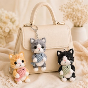 May include: Three crocheted cat keychains, each holding a small fish. One cat is orange and white with a pink fish, one is gray and white with a blue fish, and one is black and white with a green fish. All have black eyes and white whiskers.