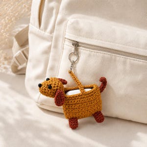 May include: A crocheted mustard yellow dog-shaped case for wireless earbuds, featuring dark red ears, feet, and a curled tail. The case has black bead eyes and a black nose, attached to a silver metal keyring clip hanging from a beige backpack.