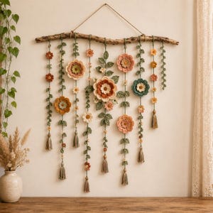 May include: A wall hanging featuring multiple crocheted flower and vine strands suspended from a natural driftwood branch. The floral elements are crafted in shades of peach, terracotta, sage green, and cream, with small tassels at the ends.