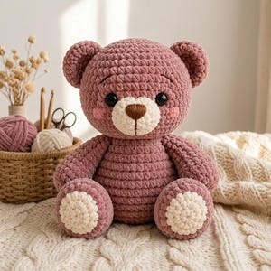 May include: A plush teddy bear made of dusty rose yarn with a cream-colored muzzle and foot pads. The bear features dark, round eyes, a brown embroidered nose, and pink blush accents on its cheeks. It has a soft, textured, crocheted appearance.