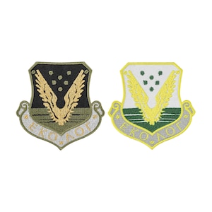 May include: Two shield-shaped embroidered patches featuring a central winged emblem above horizontal lines and five small dots. The left patch is black and olive green with gold thread, while the right is white and green with yellow thread. Both display the text EKO-ΛΟΓ.
