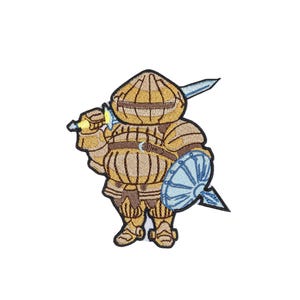 May include: An embroidered patch featuring a round, golden-armored knight figure with a bulbous helmet. The knight holds a sword over one shoulder and carries a light blue, circular shield. The design is outlined in black thread on a white background.