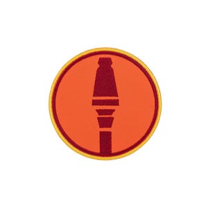 Puede incluir: Un parche bordado circular con un fondo naranja brillante y un borde rojo oscuro. Centrado en el parche hay un gráfico estilizado en rojo oscuro de un proyectil de lanzagranadas, creando un diseño audaz y de alto contraste.