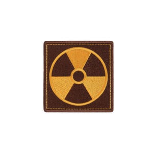 Puede incluir: Un parche bordado cuadrado con un fondo marrón oscuro y un símbolo de radiación amarillo centrado. El parche tiene un borde grueso cosido con hilo marrón oscuro a juego, creando un contorno limpio y definido para la forma cuadrada.