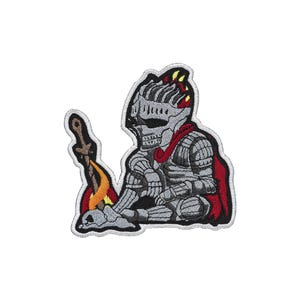 May include: An embroidered patch featuring a skeletal knight in grey armor with a red cape, sitting beside a small campfire. The knight wears a helmet and holds a sword upright in the flames, with a skull resting near the base of the fire.