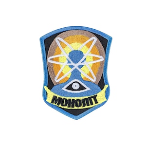 May include: A shield-shaped embroidered patch featuring a central white atom symbol over a yellow and brown circular background. Below the atom is a black eye icon on a blue field, with a yellow banner displaying the black text MOHOMT.